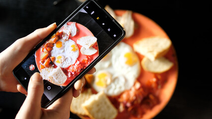 Women's hands take photos of Breakfast food using a smartphone. Fried eggs with fried potatoes, bacon and toast