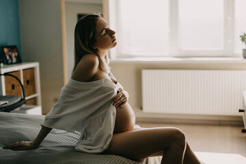 Pregnant woman in a white d at home sitting on bed with hand on belly.