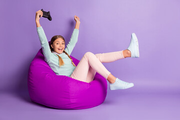 Photo of small excited lady sit beanbag hold gamepad wear green sweater pants shoes isolated violet...