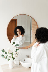 Woman with African curls in a white sweater smiles at herself in the mirror