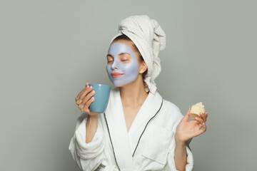Cute young woman in cosmetic face mask after bath drinking coffee on gray background