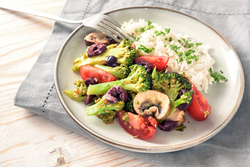 Homemade oven vegetables like broccoli, tomatoes and olives with rice on a gray plate, napkin and fork on a light wooden table