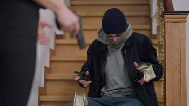 Masked scared thief taking out cash from pockets as female hand holding armed gun at front. unrecognizable Caucasian policewoman arresting burglar at crime scene. Law protection concept.