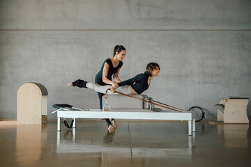 Full shot of Woman doing core exercises during Personal Pilates class on a Reformer machine, by a female instructor at a big gymnasium center, wide industrial studio. Healthy fitness lifestyle.