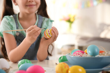 Little girl painting Easter eggs at table indoors, closeup