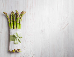 Flat lay of bunch of fresh green asparagus on light gray wooden table, copy space. The asparagus is wrapped in white paper and tied with a green satin ribbon.