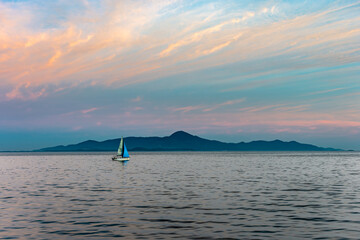 Sailboat gliding in calm sea at golden hour, multicolored sky and island on the horizon