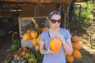 A young blonde female backpacker tourist drinks from a refreshing tropical king coconut or thambili...