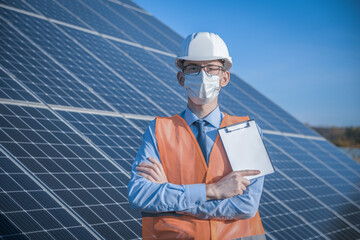 Engineer, man in uniform and mask, helmet glasses and work jacket on a background of solar panels at solar station. Technician check the maintenance.