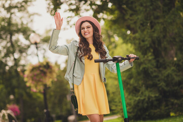 Photo of young happy smiling cheerful lovely sweet cute girl in glasses wear yellow dress hold hand say hello ride scooter in park