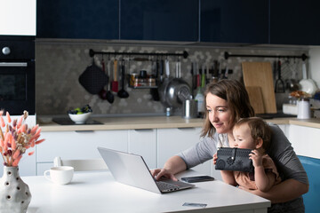 Fototapeta na wymiar Mother and her baby sitting in the kitchen with laptop. Online shopping in quarantine concept