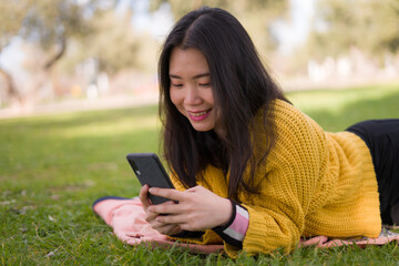 relaxed Asian woman using mobile phone in city park - lifestyle portrait of young happy and pretty Chinese girl lying on green grass networking on smart phone