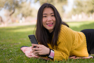 relaxed Asian woman using mobile phone in city park - lifestyle portrait of young happy and pretty Japanese girl lying on green grass networking on smart phone