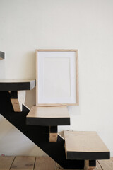 Closeup of square empty wooden picture frame on wooden stairs. White wall background. Scandinavian interior, home design. Art concept. Artistic mockup scene. Front view