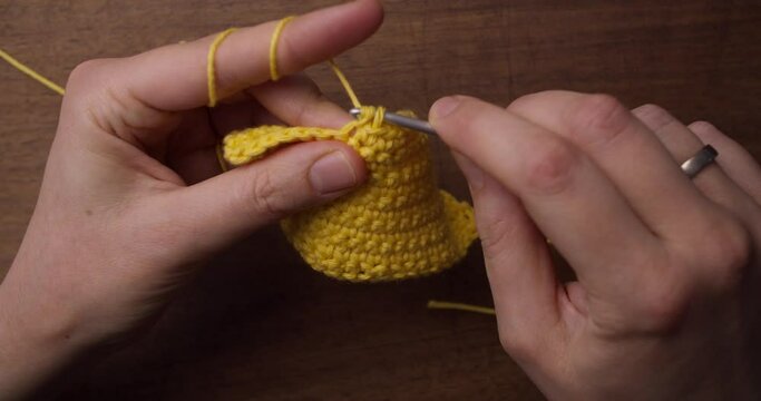 Crochet with yellow wool. Caucasian hands handling a crochet needle and making a yellow scarf. Static pov top shot with soft light from the left.