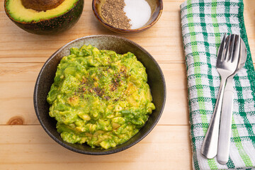 Fresh guacamole on a dish placed on a wooden table with ingredients for homemade guacamole: avocados, lemon, salt, and pepper. Top view.  Concept of traditional Mexican preparation