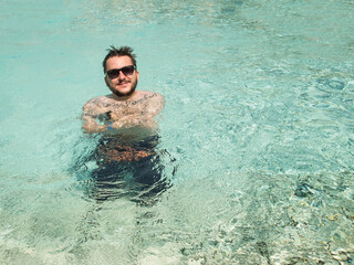 Portrait of smiling bearded tattooed young adult man wearing sunglasses at swimming pool