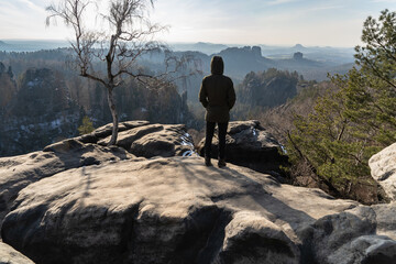 A man standing up on a huge rock at a mountain top and looking down to the slightly hazy valley...