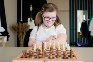 Portrait of young smiling girl with Down syndrome playing chess on the game board. Disabled kids...