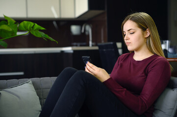 Obraz premium Young woman is sitting on the coach holding cellphone on her hands