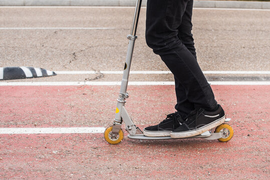 Detalle de un hombre joven vestido con ropa negra de invierno montando en patinete por un carril bici de la ciudad. Scooter no el&eacute;ctrico. Concepto de transporte sostenible