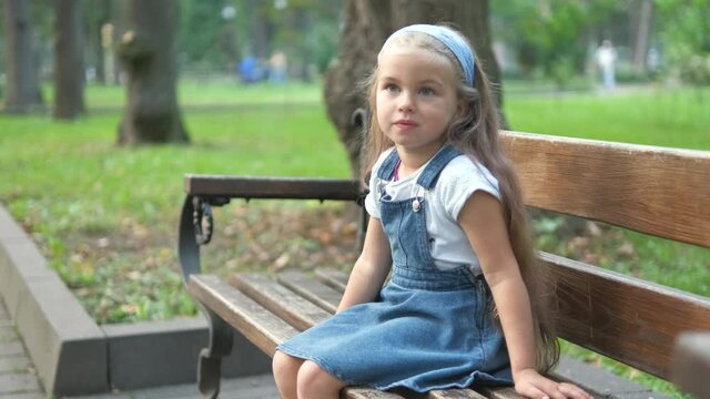 Little child girl sitting alone on a bench in summer park.