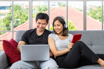 Young Asian couple relaxing sitting on sofa using laptop computer and digital tablet working from home