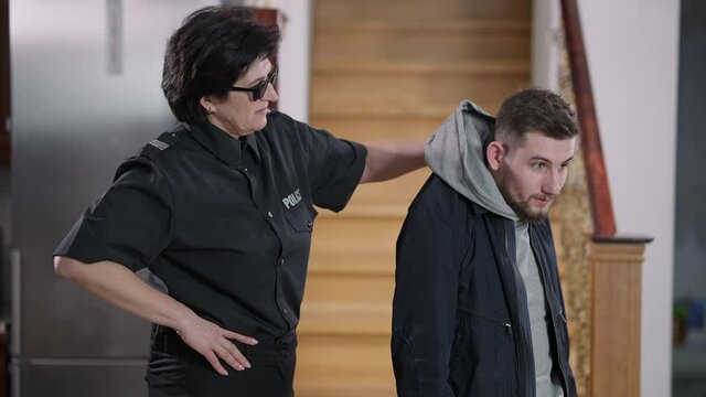 Scared young Caucasian thief grabbed by the collar. Strong confident female police officer catching bandit robbing house indoors. Policewoman in sunglasses arresting burglar. Law protection.