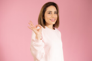 Young beautiful woman wearing casual sweater standing over pink isolated background smiling positive doing okay sign with hand and fingers. Successful expression.