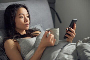 Fototapeta premium Happy beautiful asian woman is relaxing at bed while using smartphone at the morning