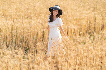 Naklejka na ściany i meble Happy young beautiful woman wearing black hat and white dress enjoying herself walking in the golden barley filed on a late afternoon