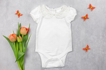 White baby girl bodysuit mockup on the gray concrete background with tulips flowers and spring decoration. Design onesie template, print presentation mock up. Top view. Flat Lay.