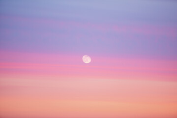 Obraz premium Beautiful backdrop of sunset sky of pink orange lilac colors and moon in center. Colorful smooth dawn sky gradient. Nature background of sunrise. Morning heaven. Slightly cloudy evening atmosphere.