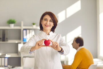 Friendly female cardiologist holding a small red heart as a symbol of a healthy important human organ. Concept of health, cardiology and prevention of heart disease. Blurred background.