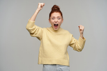 Excited girl with ginger hair gathered in a bun. Wearing pastel yellow sweater and jeans. Clench and lifts her fists. Scream and celebrate victory. Watching at the camera isolated over grey background