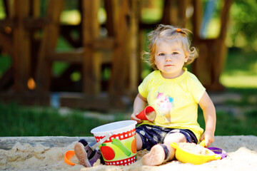 Cute toddler girl playing in sand on outdoor playground. Beautiful baby having fun on sunny warm summer sunny day. Happy healthy child with sand toys and in colorful fashion clothes.