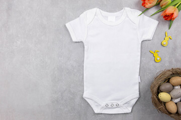 White baby girl or boy bodysuit mockup on the gray concrete background with tulips flowers and easter decoration. Design onesie template, print presentation mock up. Top view. Flat Lay. Copy space.