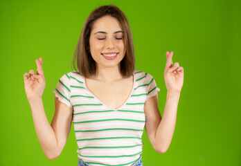 Portrait of a smiling young woman standing over green background, holding fingers crossed for good luck