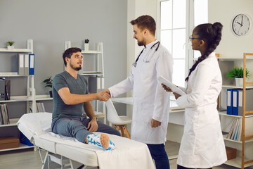 Obraz premium Thankful patient with foot fracture and doctor handshaking after successful trauma treatment. Grateful man with bandage on broken ankle and interracial clinic staff team at ward. Medicine, healthcare