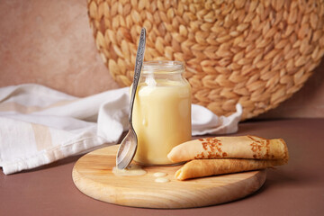 Jar with sweet condensed milk and thin pancakes on color background