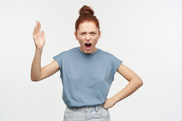 Teenage, angry looking woman with ginger hair gathered in a bun. Wearing blue t-shirt and jeans. Raise her hand and put another on a hip, shout. Watching at the camera isolated over white background