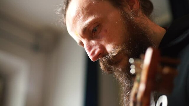 The focus of the musician's attention is on creating a melody. He looks down, feels harmony. He touches the strings with his hands. Bearded face.