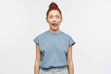 Shocked looking woman, beautiful girl with ginger hair gathered in a bun. Wearing blue t-shirt and jeans. Lick her lip, shows tongue. Watching at the camera isolated over white background