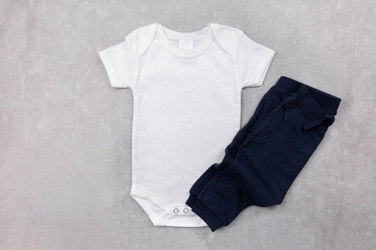 White Baby Girl Or Boy Bodysuit Mockup On The Gray Concrete Background With Dark Blue Panties. Design Onesie Template, Print Presentation Mock Up. Top View. Flat Lay.