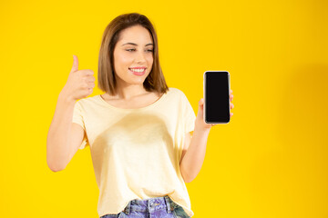 Image of pretty young woman isolated over yellow background. Looking camera showing display of mobile phone.