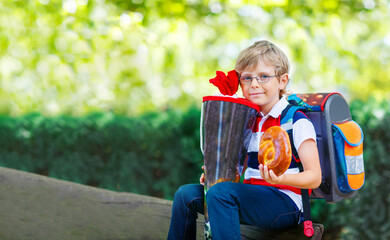 Fototapeta premium Happy little kid boy with glasses and backpack or satchel on his first day to school . Child outdoors on warm sunny day, Back to school concept. Kid with traditional schoolbag in German Schultuete.