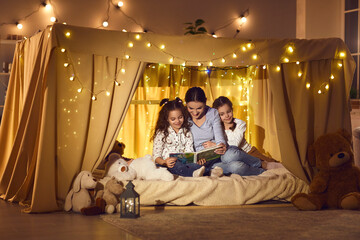 Obraz na płótnie Canvas Happy family enjoying a good quiet evening at home. Mommy telling fairy tales to children. Young mother and two little daughters reading book of bedtime stories sitting in cozy playroom tent together