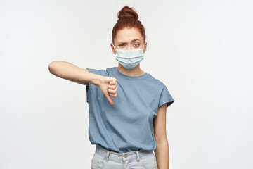 Portrait of disappointed redhead girl with hair gathered in a bun. Wearing blue t-shirt and protective mask. Showing thumb down in disagreement. Watching at the camera isolated over white background