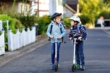Two school kid boys in safety helmet riding with scooter in the city with backpack on sunny day. Happy children in colorful clothes biking on way to school. © Irina Schmidt