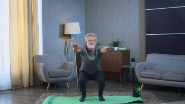 Portrait of a gray-haired senior man with a beard squats. Does home exercises. Grandfather in excellent athletic body shape. Old man in sportswear. Squatting for back health in old age.